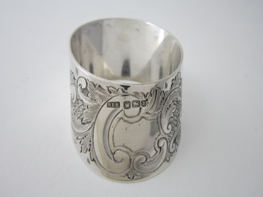 A Victorian sterling silver napkin ring featuring intricate foliate scroll engraving and visible British hallmarks on a reflective curved surface. View III