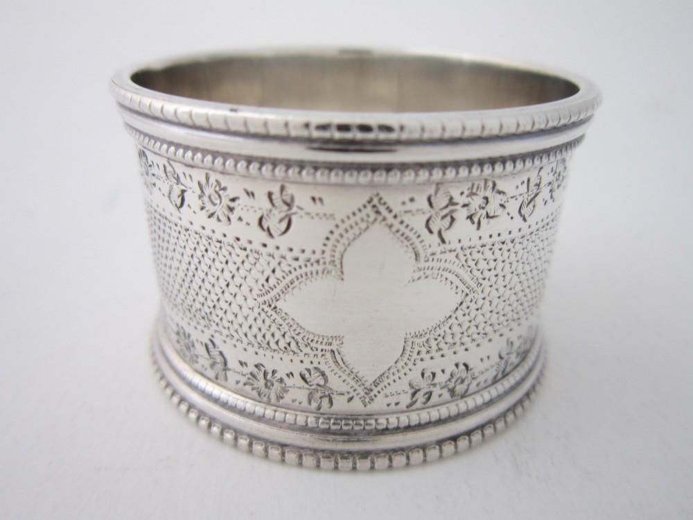 An antique sterling silver napkin ring featuring intricate bright-cut floral engraving, engine-turned lattice patterning, a central cartouche, and beaded borders.