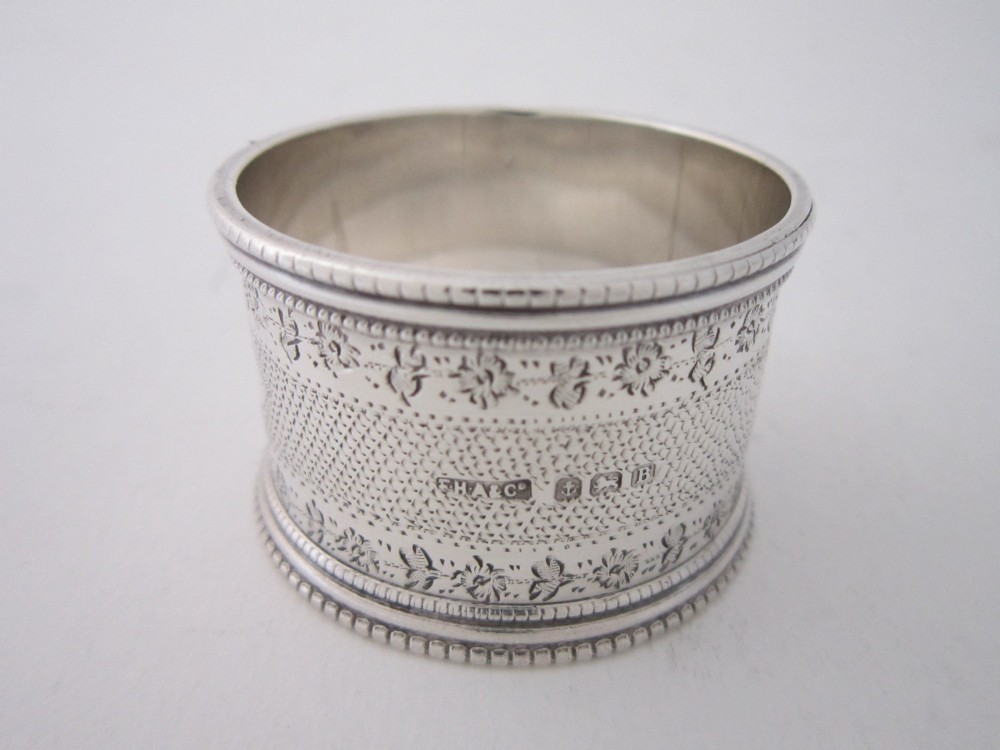 An antique sterling silver napkin ring featuring intricate bright-cut floral engraving, engine-turned lattice patterning, a central cartouche, and beaded borders. View II