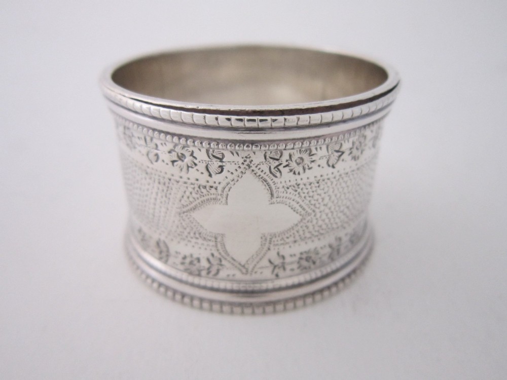 An antique sterling silver napkin ring featuring intricate bright-cut floral engraving, engine-turned lattice patterning, a central cartouche, and beaded borders. View III