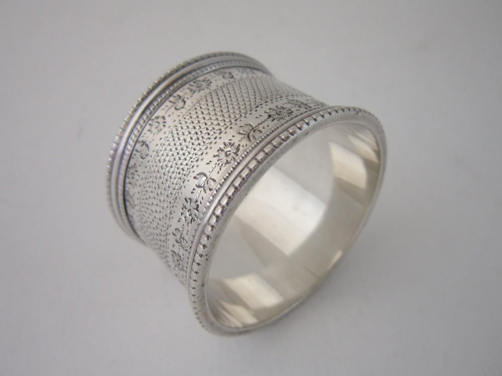 An antique sterling silver napkin ring featuring intricate bright-cut floral engraving, engine-turned lattice patterning, a central cartouche, and beaded borders. View IV