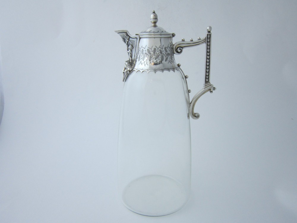 A Victorian clear glass claret jug with an engraved sterling silver collar, hinged lid with finial, and an ornate scroll handle.