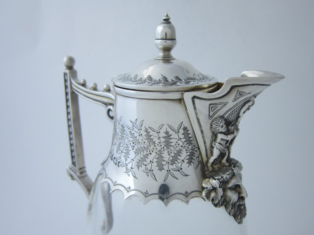 A Victorian clear glass claret jug with an engraved sterling silver collar, hinged lid with finial, and an ornate scroll handle. View V