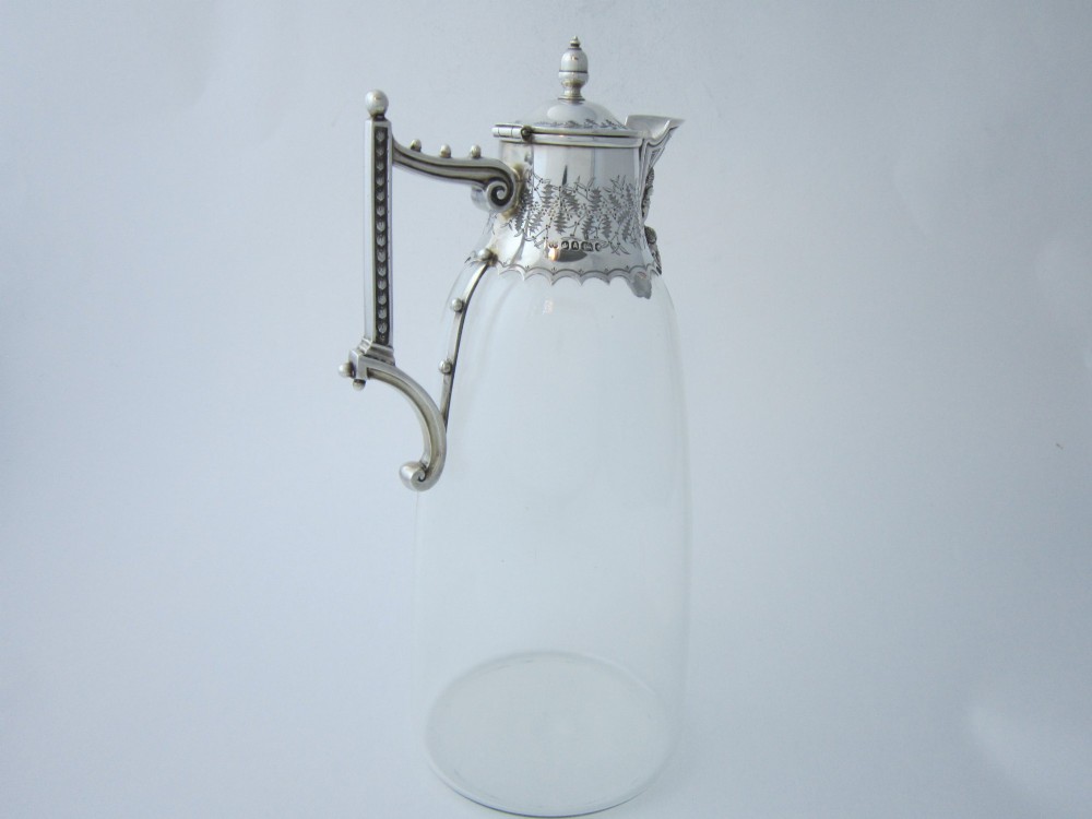 A Victorian clear glass claret jug with an engraved sterling silver collar, hinged lid with finial, and an ornate scroll handle. View VI