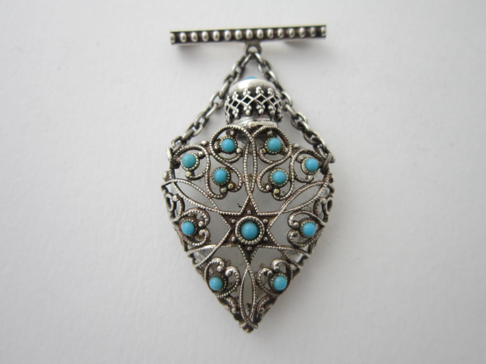 A 1920s Continental silver scent bottle featuring clear glass enclosed in silver filigree overlay set with turquoise cabochons, suspended from a beaded bar pin.