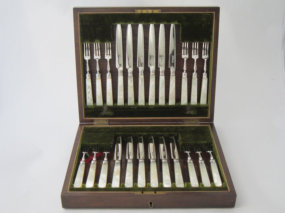 An open fitted wooden canteen displaying a set of sterling silver dessert knives and forks with mother-of-pearl handles set against a green velvet lining.