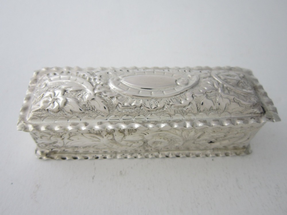 A small rectangular Victorian sterling silver trinket box featuring embossed floral scrolls, scalloped edges, and a blank central cartouche on the hinged lid.