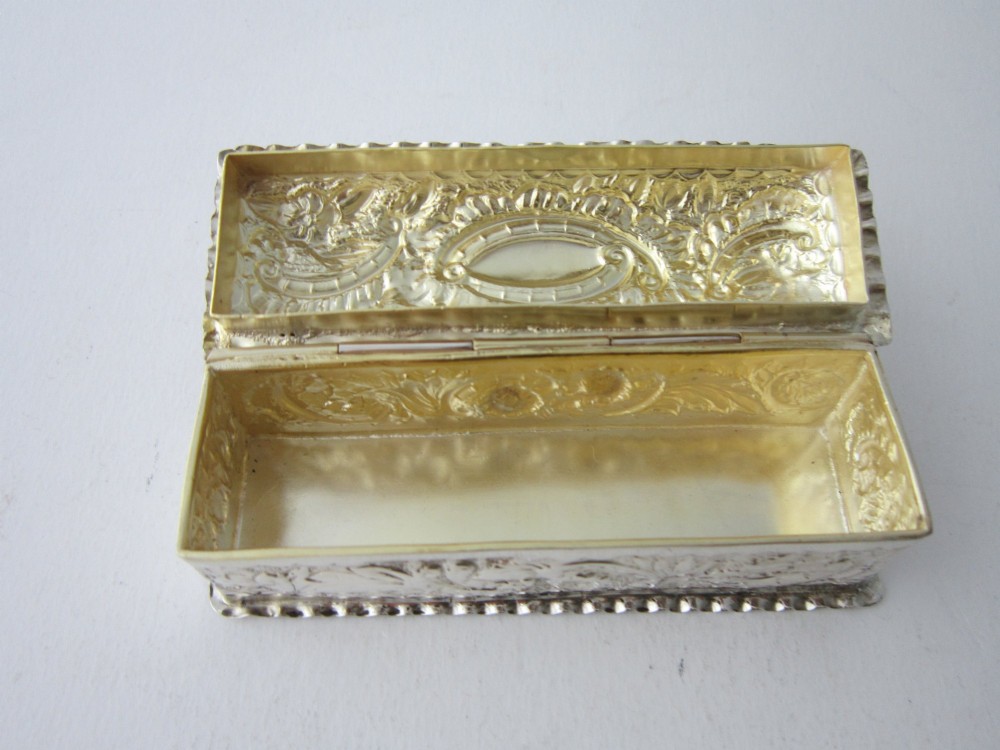 A small rectangular Victorian sterling silver trinket box featuring embossed floral scrolls, scalloped edges, and a blank central cartouche on the hinged lid. View VI