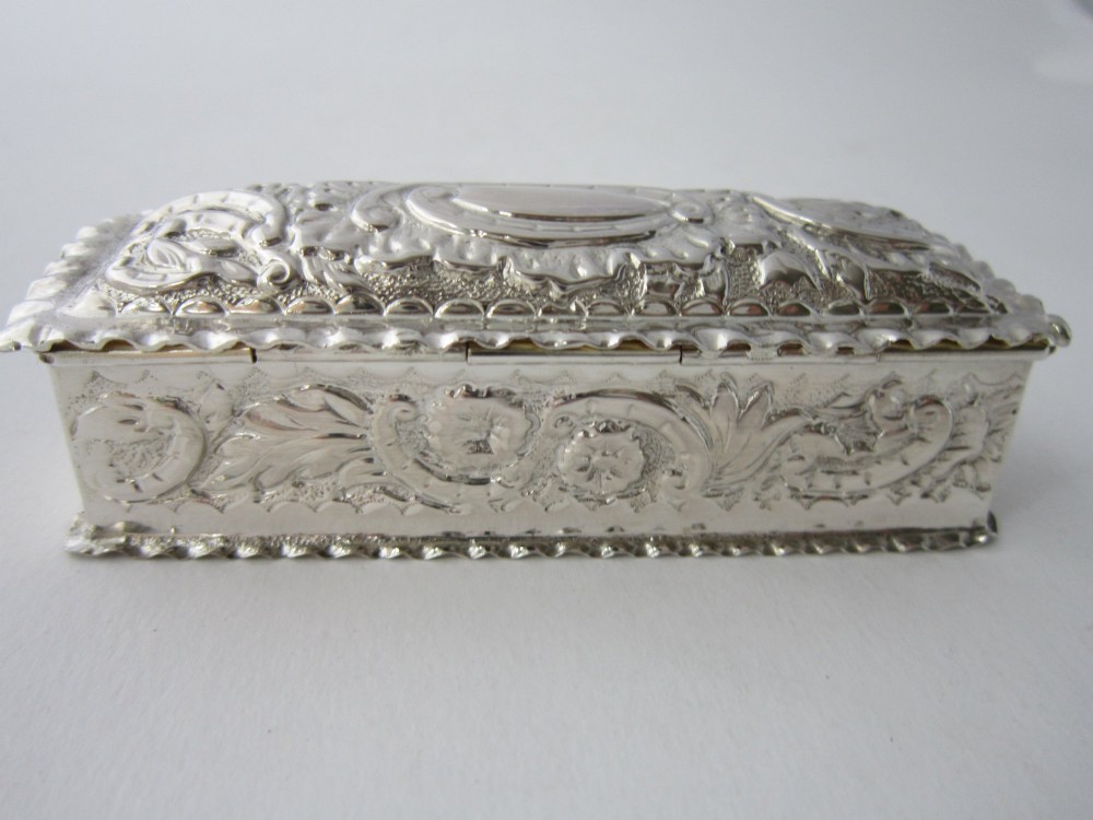 A small rectangular Victorian sterling silver trinket box featuring embossed floral scrolls, scalloped edges, and a blank central cartouche on the hinged lid. View VIII