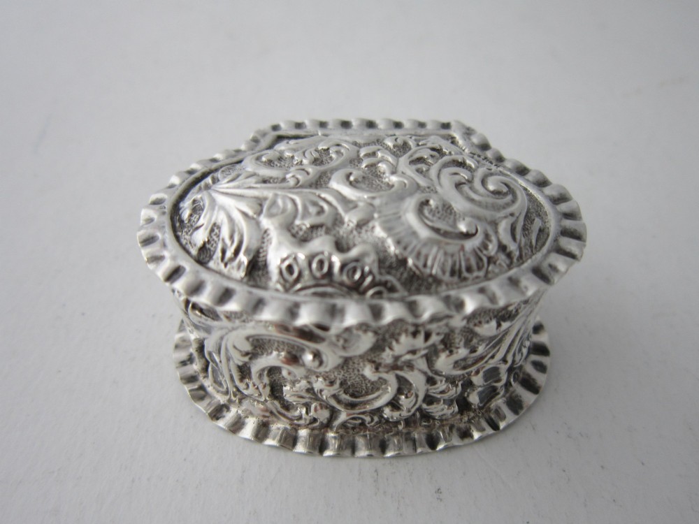 An oval Victorian sterling silver pill box decorated with embossed scrollwork and crimped fluted rims on the lid and base.