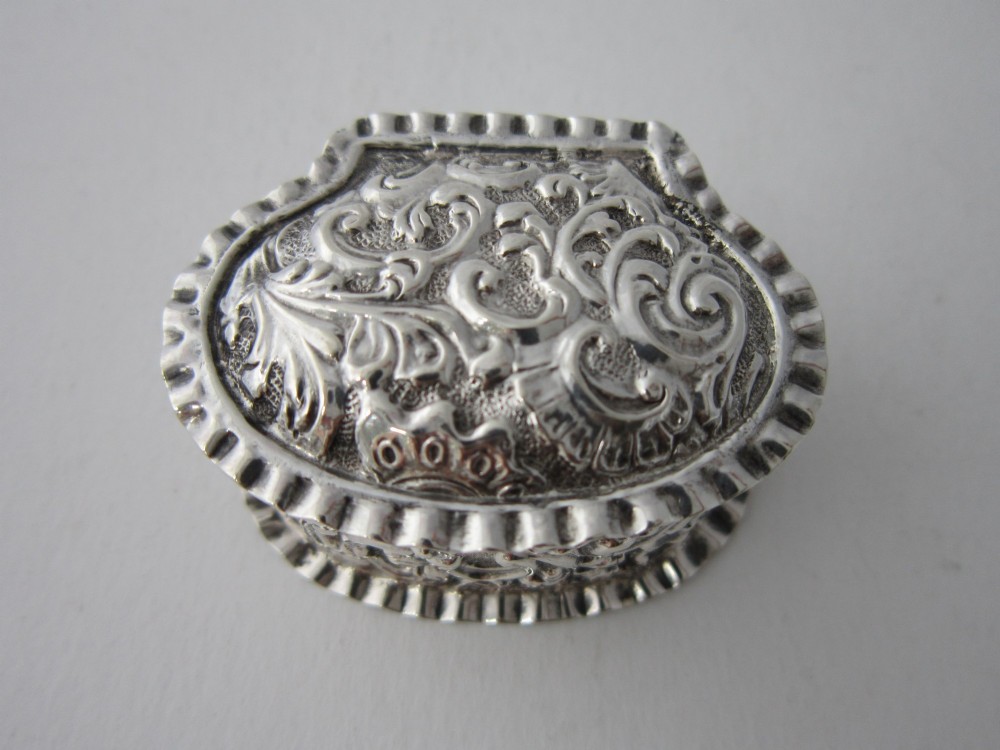 An oval Victorian sterling silver pill box decorated with embossed scrollwork and crimped fluted rims on the lid and base. View IV