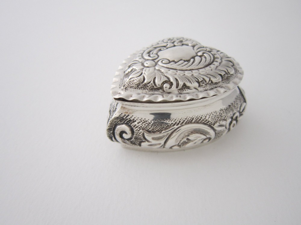 A small heart-shaped sterling silver trinket box featuring intricate repousse scrollwork, floral motifs, a plain central cartouche, and a crimped rim. View II