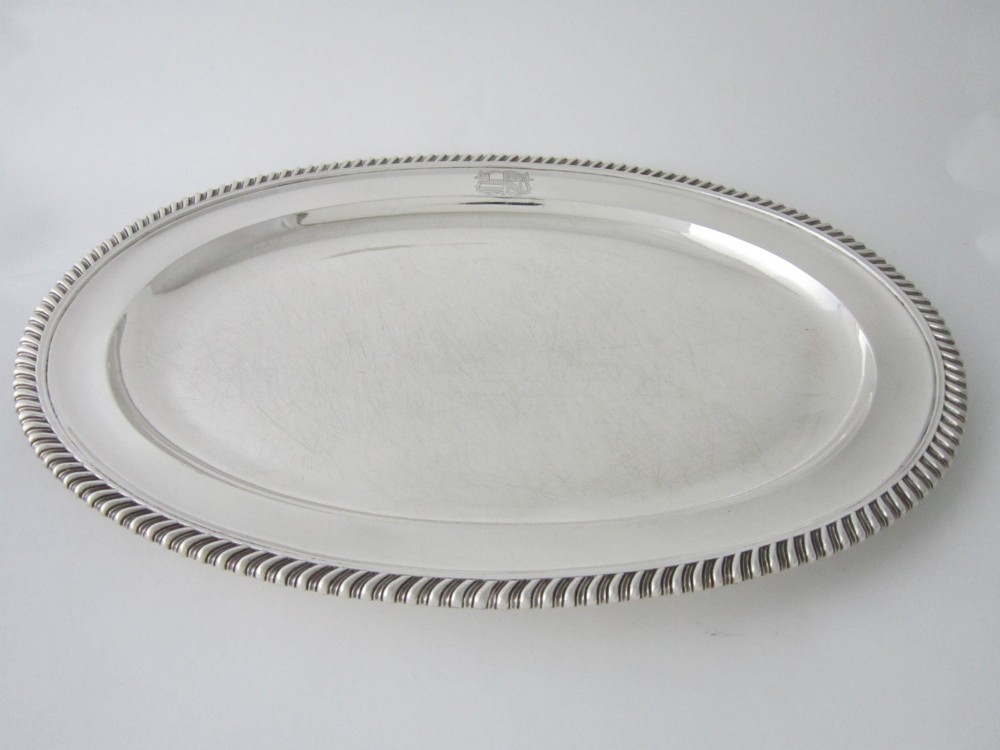 An oval William IV sterling silver meat platter featuring a decorative gadrooned border and an engraved coat of arms on the upper rim.