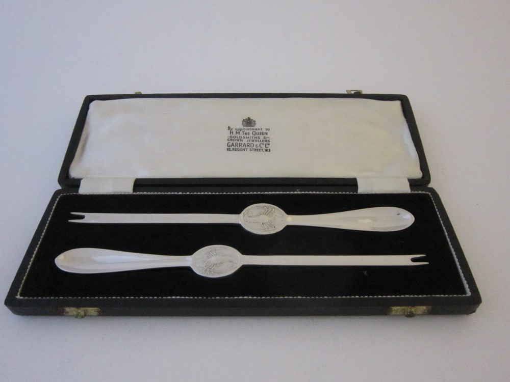 A pair of sterling silver lobster picks with embossed crustacean central medallions displayed inside a fitted black satin and velvet presentation box.