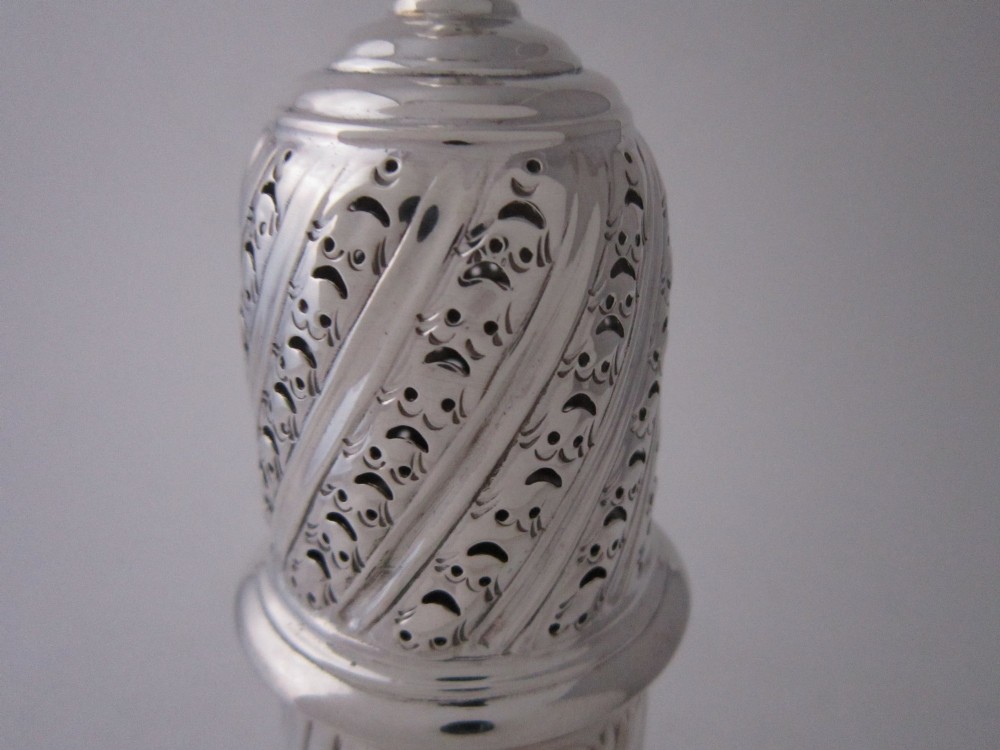 A Victorian sterling silver sugar caster featuring a spirally fluted pierced dome lid, a flame finial, and embossed cartouche detailing on a pedestal foot. View V