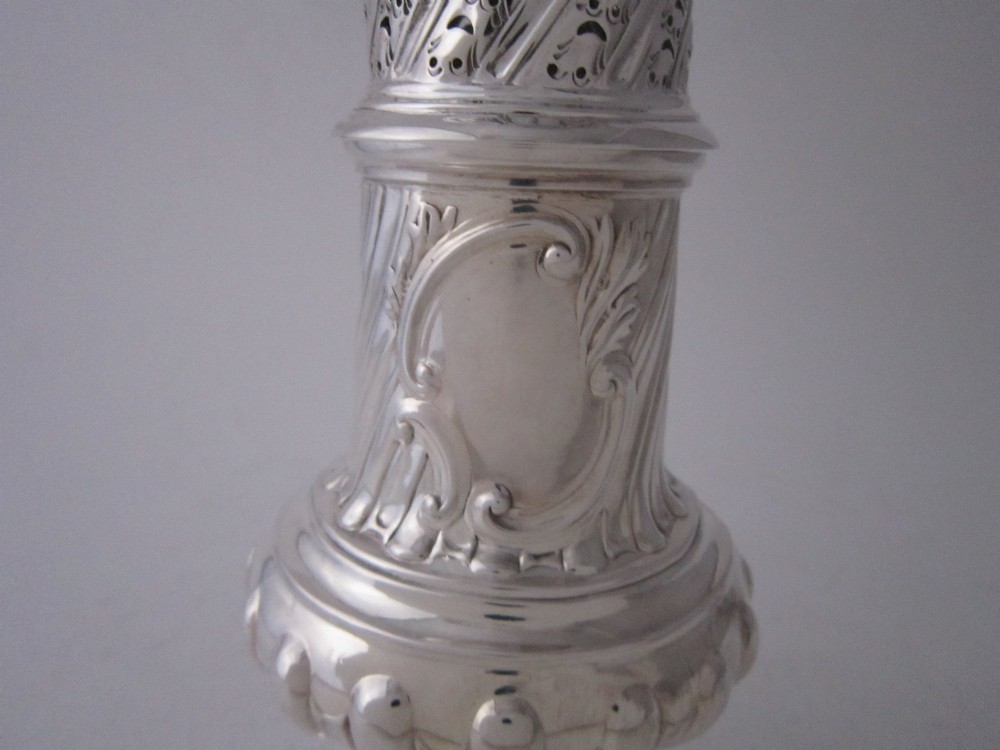 A Victorian sterling silver sugar caster featuring a spirally fluted pierced dome lid, a flame finial, and embossed cartouche detailing on a pedestal foot. View VI