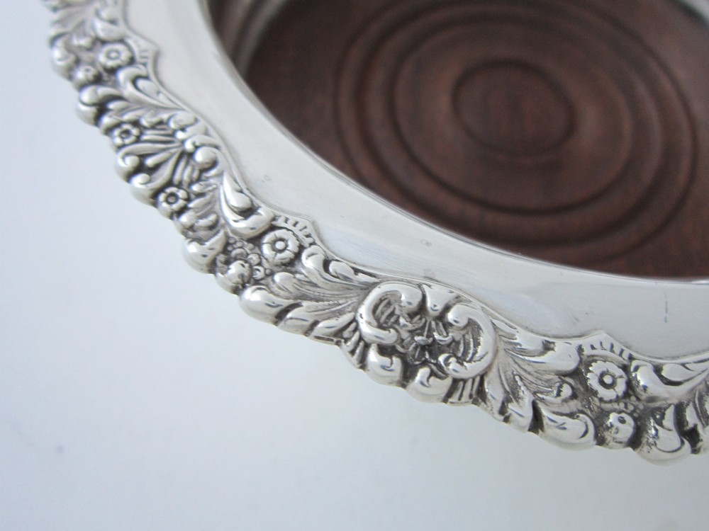 A circular George V sterling silver wine coaster featuring an ornate floral scroll rim and a polished stepped body with a wood insert base. View III