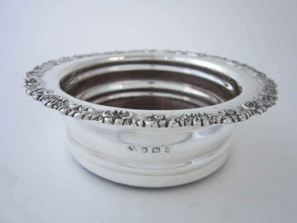 A circular George V sterling silver wine coaster featuring an ornate floral scroll rim and a polished stepped body with a wood insert base. View V
