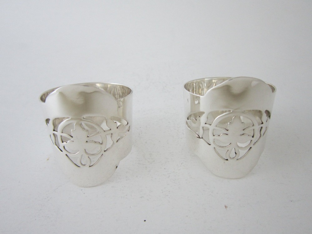 A pair of polished sterling silver napkin rings featuring pierced fretwork crest designs and shaped borders.