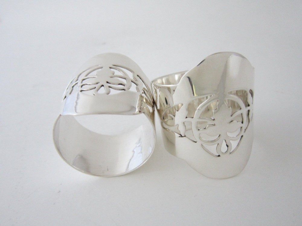 A pair of polished sterling silver napkin rings featuring pierced fretwork crest designs and shaped borders. View IV