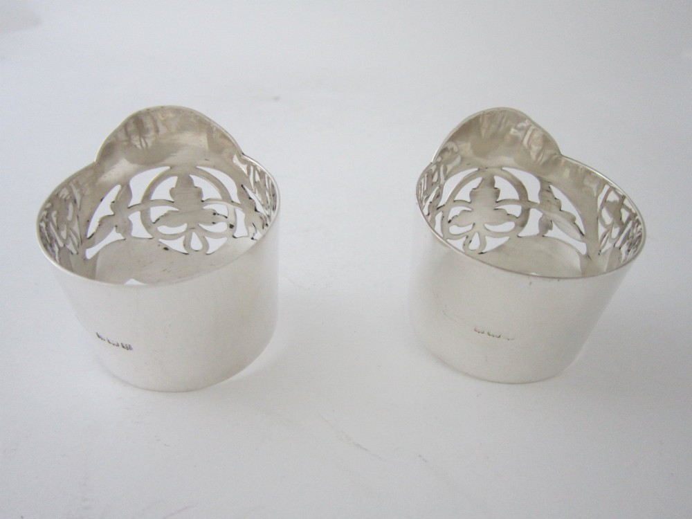 A pair of polished sterling silver napkin rings featuring pierced fretwork crest designs and shaped borders. View V