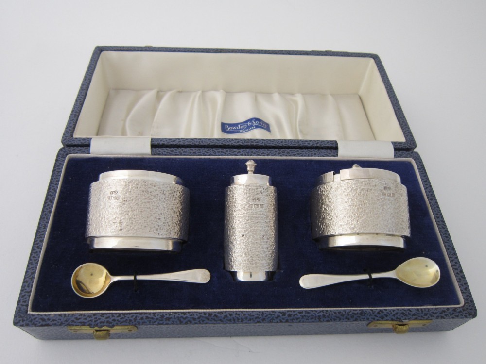 A three-piece Elizabeth II sterling silver condiment set with textured bark-effect bodies, including a mustard pot, salt pot, pepper pot, and two small spoons, housed in a blue presentation case.