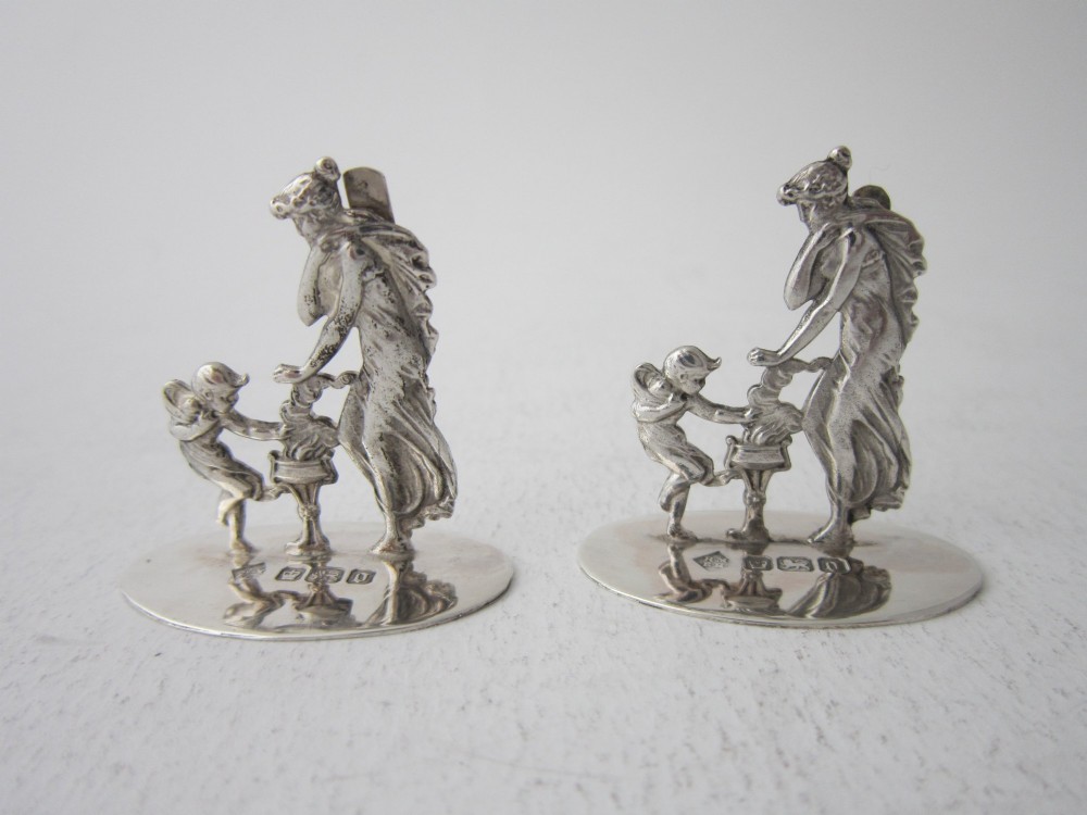 A pair of Edwardian sterling silver place card holders featuring classical female figures and cherubs on round hallmarked bases.