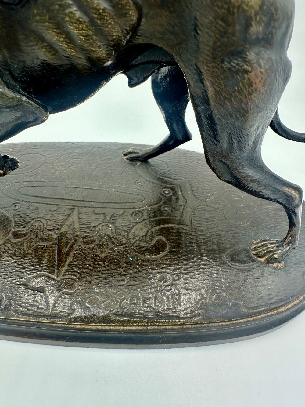 A dark patinated bronze sculpture depicting a greyhound scratching its neck, set on an oval decorated base. View V