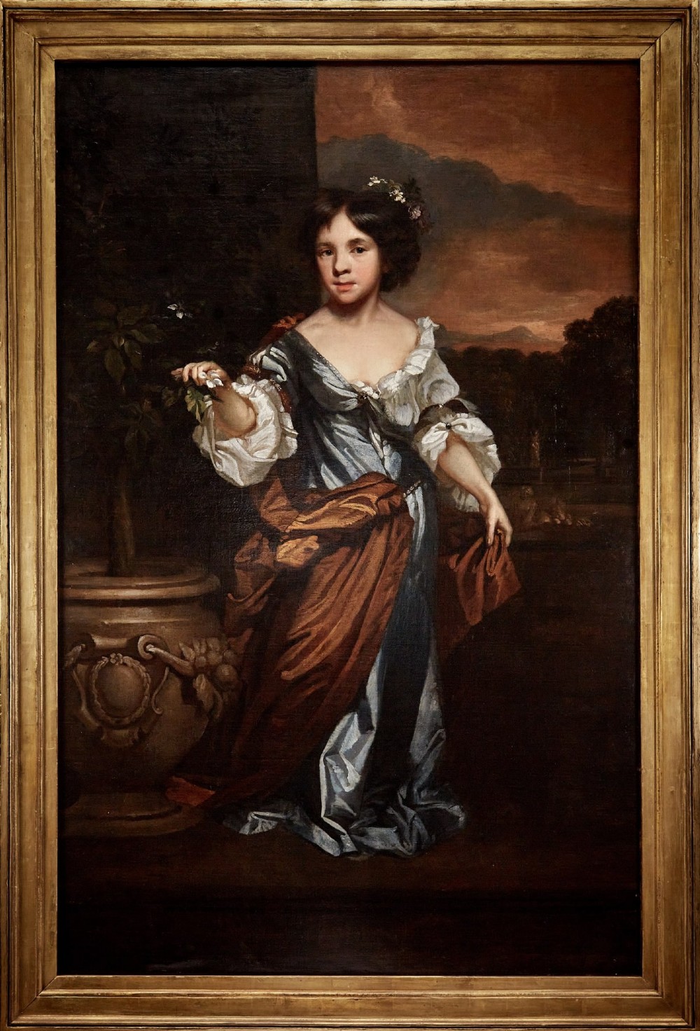 A full-length 17th-century oil portrait of a young girl wearing a blue gown standing on a terrace beside a stone urn inside a gilt wood frame.
