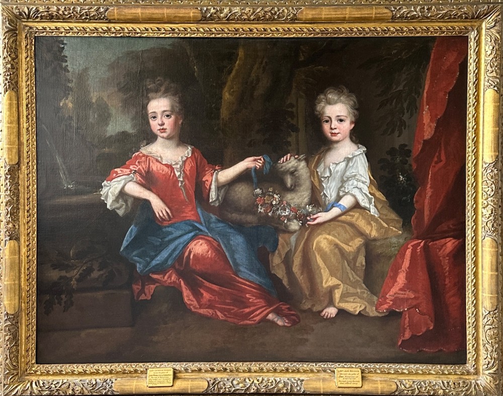An 18th-century oil painting depicting two young girls seated in a natural landscape with a lamb, presented within an ornate carved giltwood frame.