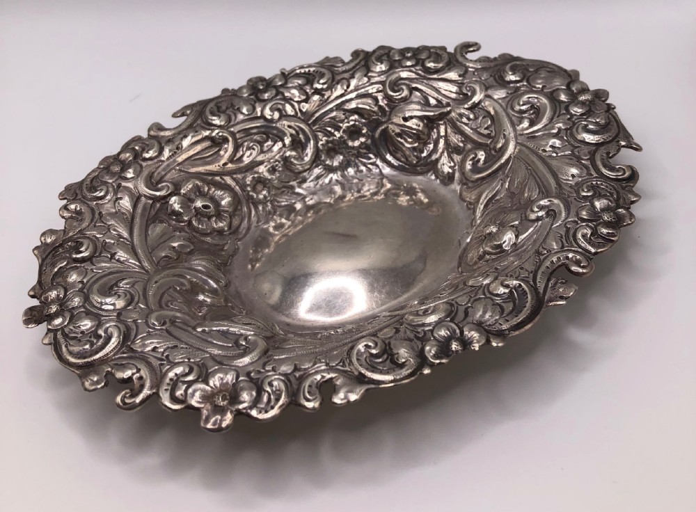 An oval Victorian sterling silver dish with detailed repousse floral motifs and a scalloped scrolling edge.