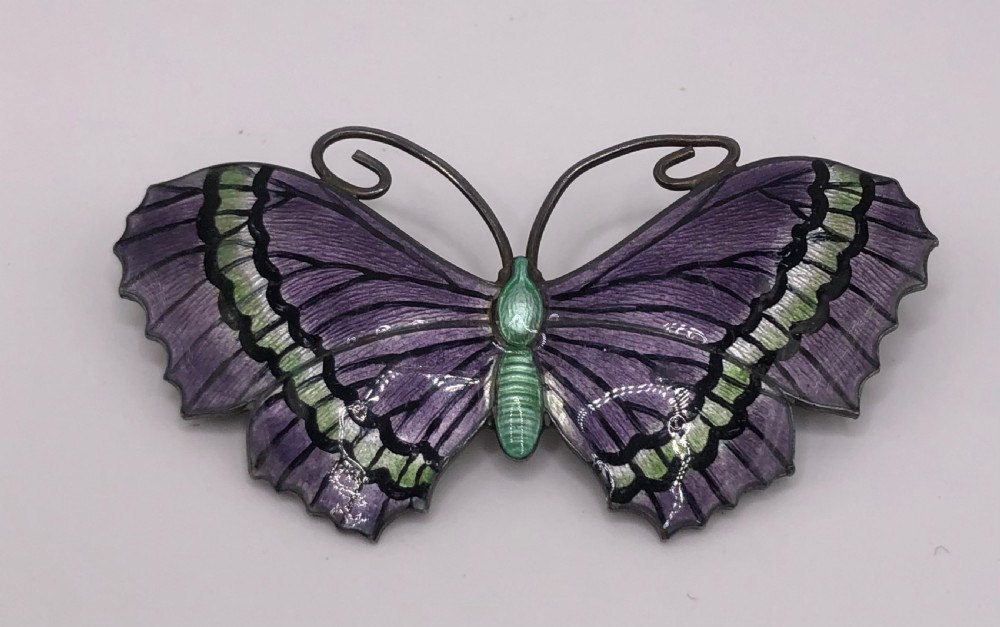 A silver butterfly brooch featuring purple and green guilloche enamel wings with black details and coiled wire antennae.