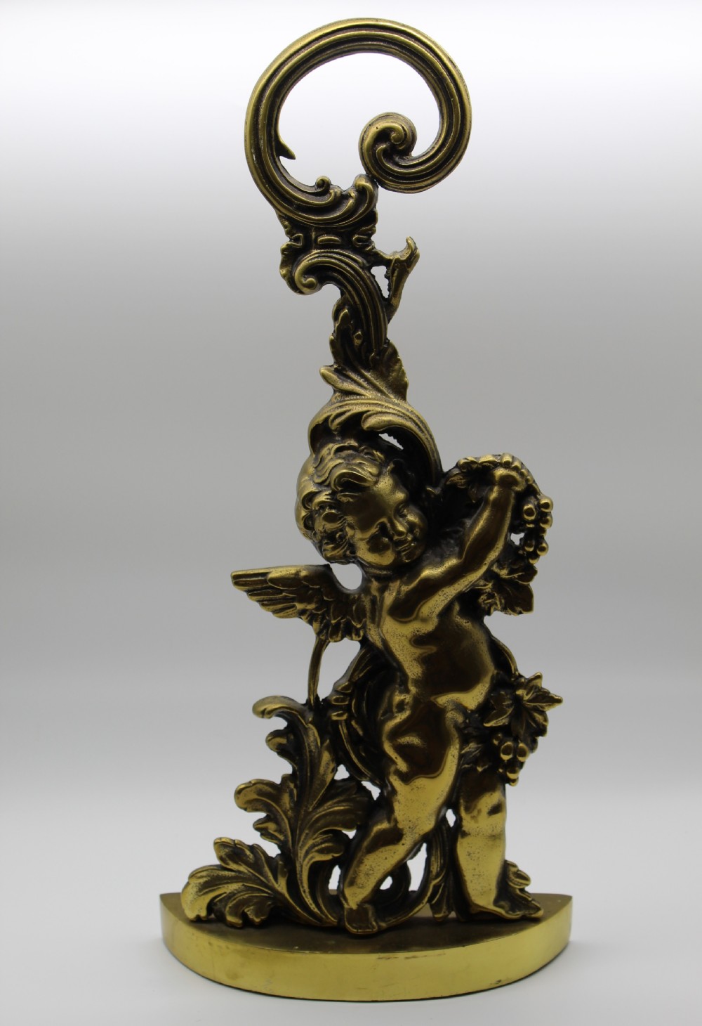 A Victorian cast brass doorstop featuring a standing winged cherub amidst acanthus leaf scrolls topped with a curved carrying handle.