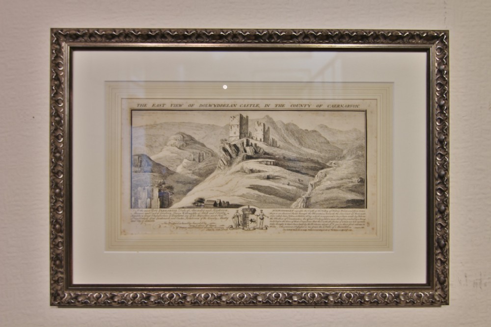 An 18th-century monochrome framed engraving depicting Dolwyddelan Castle perched on a Welsh hillside with text and coat of arms at the bottom.