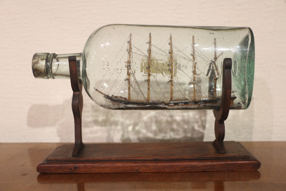 A nineteenth-century glass bottle containing a detailed miniature sailing ship model, resting on a carved wooden display stand.