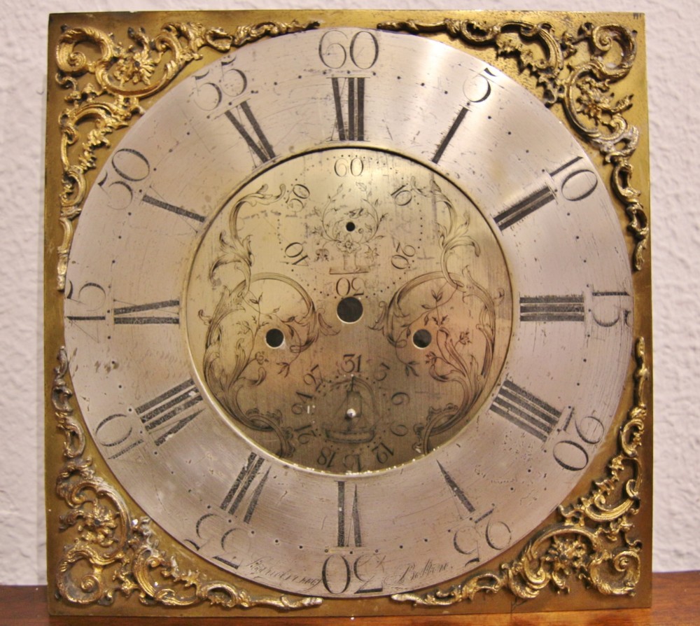 A square 18th-century brass longcase clock dial featuring an engraved silvered chapter ring, ornate cast brass corner spandrels, and central date and seconds apertures.