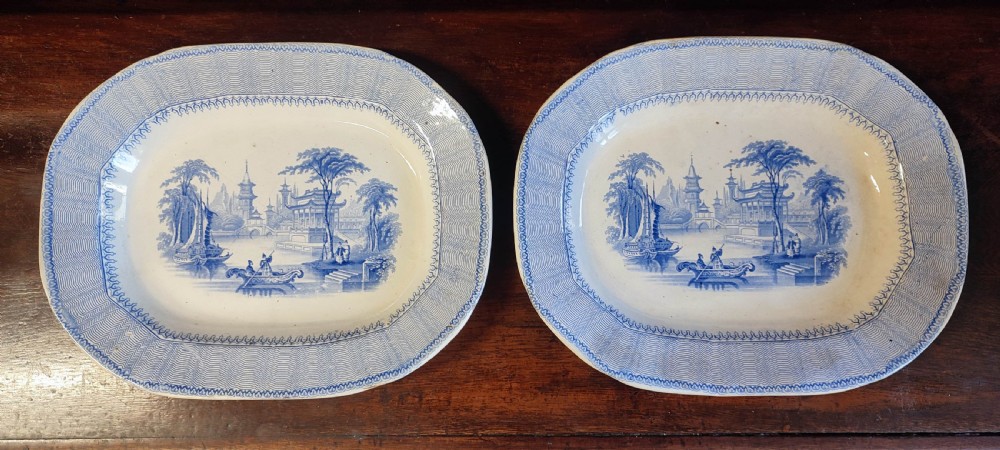 A pair of 19th-century oval earthenware meat dishes featuring blue transfer-printed chinoiserie landscape scenes with pagodas, boats, and detailed geometric borders.