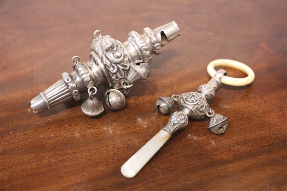 A pair of 18th-century silver childs rattles featuring intricate repousse scrollwork, attached bells, an integral whistle, and a mother-of-pearl handle.