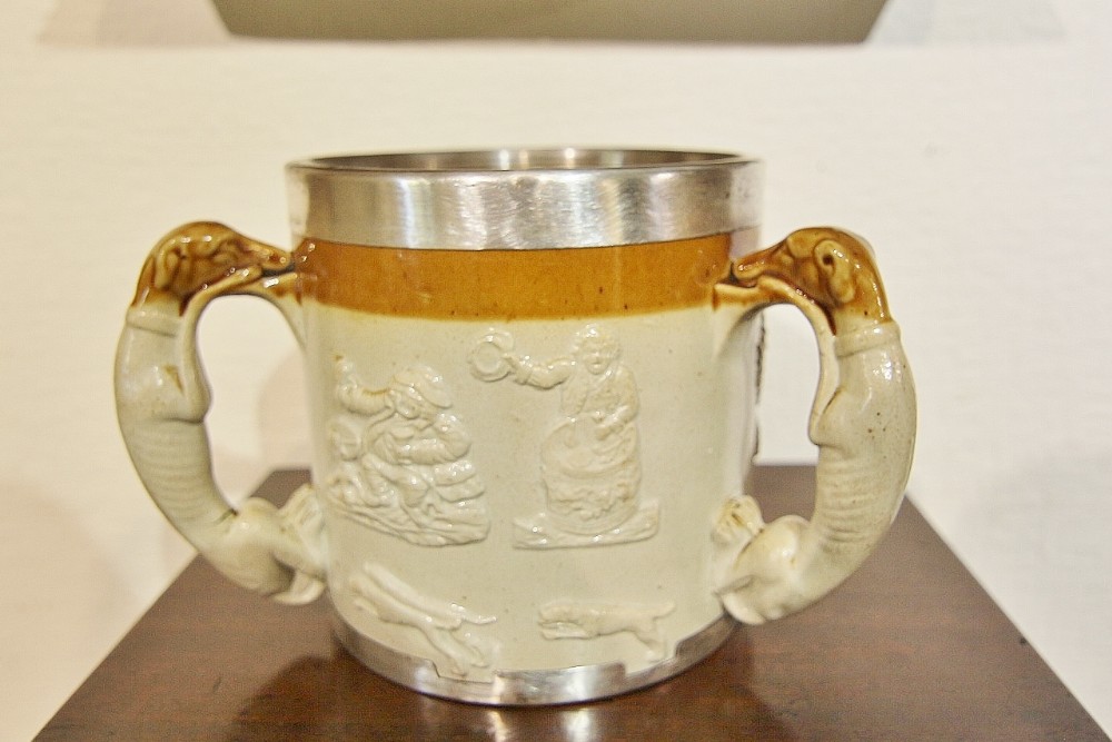 A 19th-century two-handled stoneware harvest cup featuring a silver-rimmed border, greyhound handles, and sprigged relief figures of hounds and drinkers against a buff and ochre background.