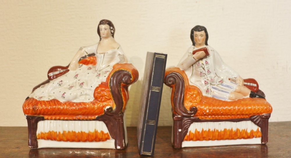 A pair of Staffordshire earthenware bookends featuring hand-painted male and female figures reclining on orange and brown scroll-arm chaises.