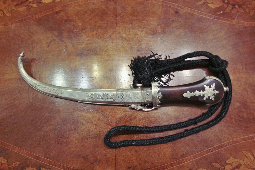 A traditional Moroccan Berber Koummya dagger featuring an engraved silvered scabbard, carved wooden hilt with metal overlays, and an attached black cord braid with tassels.