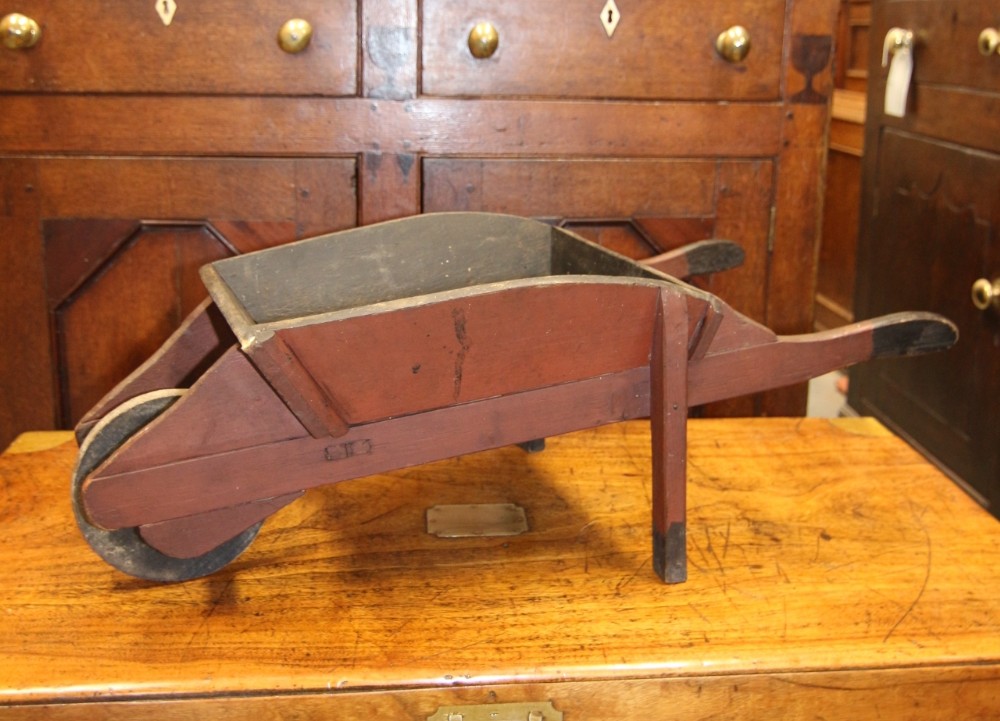 A small antique Welsh folk art wooden wheelbarrow finished in terracotta red paint with a solid wooden wheel and long handles.