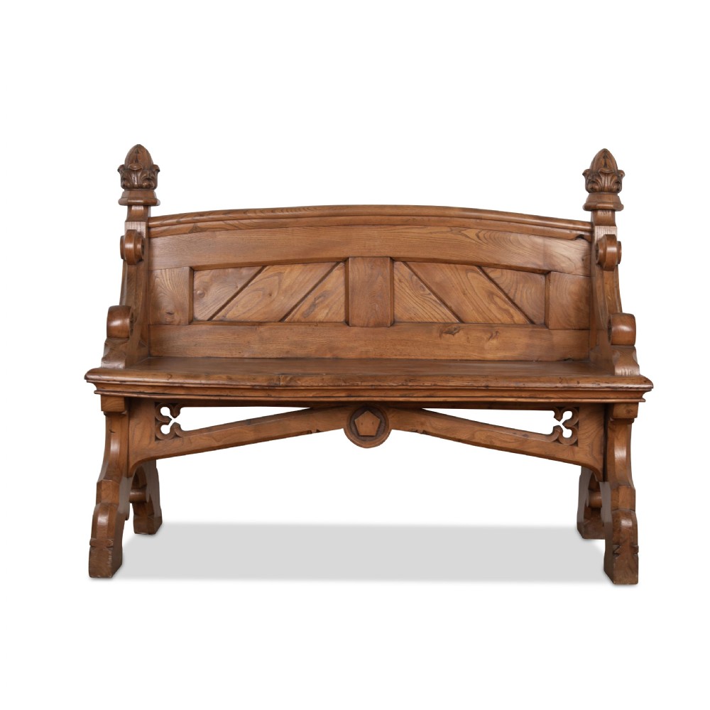 A 19th-century carved elm bench featuring pointed finial posts, a geometric panelled backrest, and trefoil cut-outs on the lower frame.