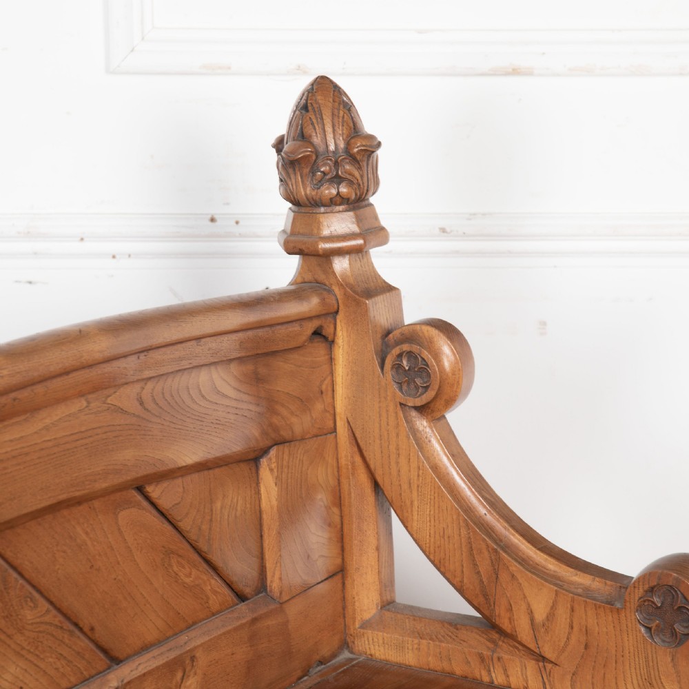 A 19th-century carved elm bench featuring pointed finial posts, a geometric panelled backrest, and trefoil cut-outs on the lower frame. View VII