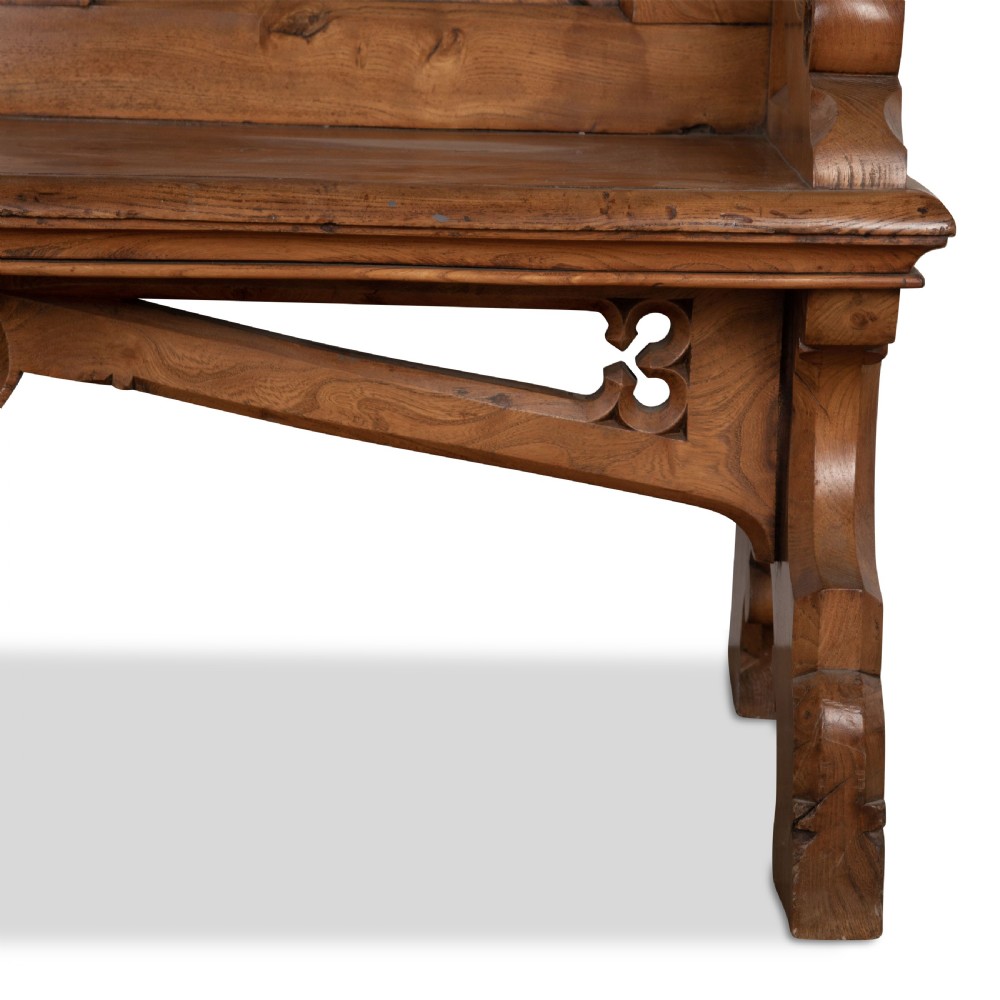 A 19th-century carved elm bench featuring pointed finial posts, a geometric panelled backrest, and trefoil cut-outs on the lower frame. View X
