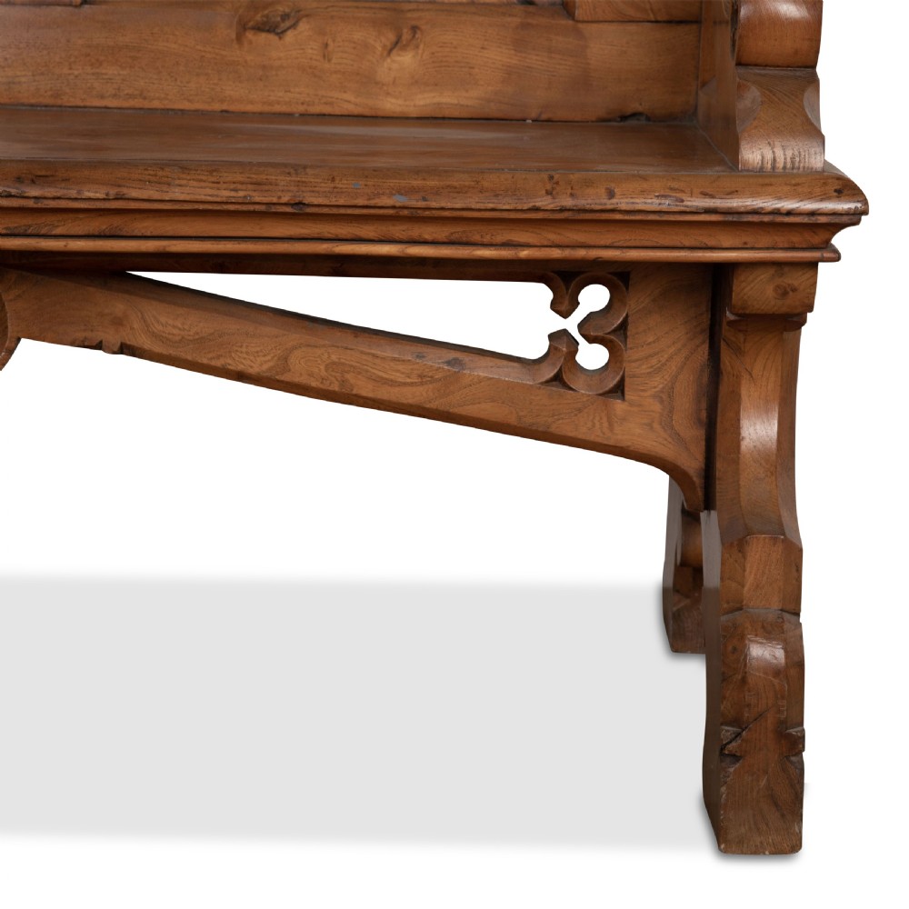 A 19th-century carved elm bench featuring pointed finial posts, a geometric panelled backrest, and trefoil cut-outs on the lower frame. View XIV