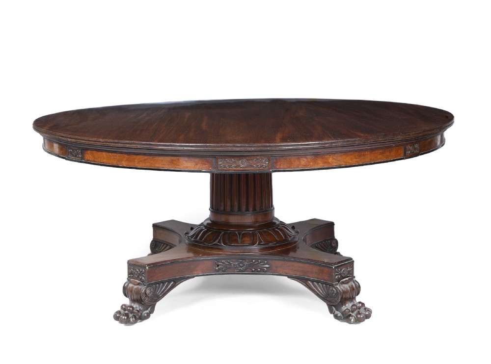 A circular Regency mahogany centre table featuring a carved frieze, a fluted pedestal column, and a concave four-legged plinth base with carved feet.