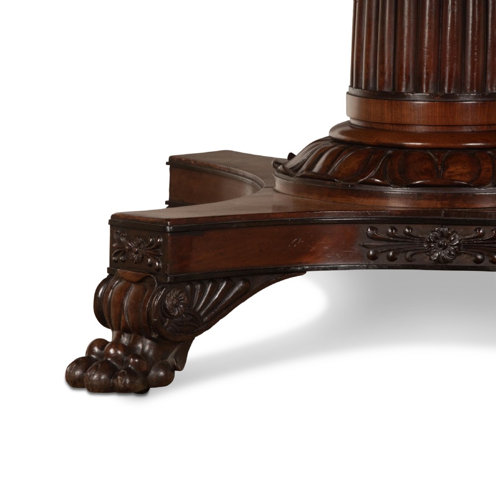 A circular Regency mahogany centre table featuring a carved frieze, a fluted pedestal column, and a concave four-legged plinth base with carved feet. View IX