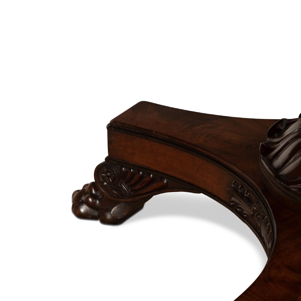 A circular Regency mahogany centre table featuring a carved frieze, a fluted pedestal column, and a concave four-legged plinth base with carved feet. View X