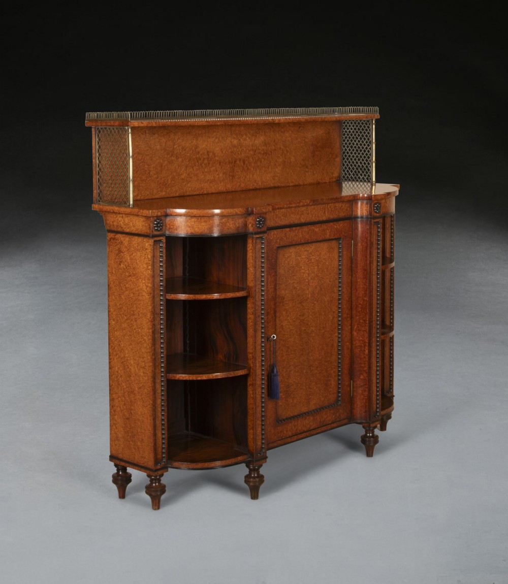 A Regency amboyna wood side cabinet featuring open curved side shelves, a central panel door, turned feet, and an upper brass gallery.